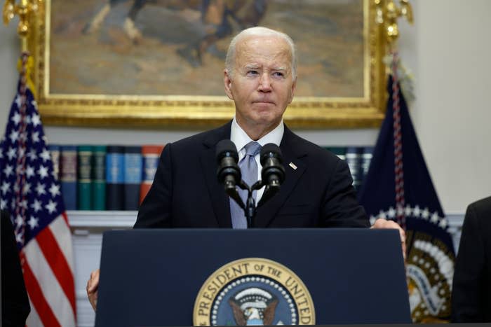 President Joe Biden speaking at a podium with the presidential seal, flanked by U.S. and presidential flags, a painting, and bookshelves in the background