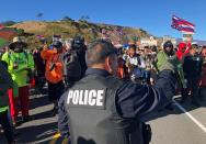 A police officer gestures at demonstrators blocking a road at the base of Hawaii's tallest mountain, Monday, July 15, 2019, in Mauna Kea, Hawaii, who are protesting the construction of a giant telescope on land that some Native Hawaiians consider sacred. (AP Photo/Caleb Jones)