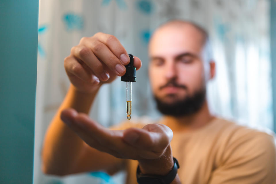 In the background, a man with a beard and moustache holds a dropper with beard oil in it in the foreground and is about to squeeze some onto his palm