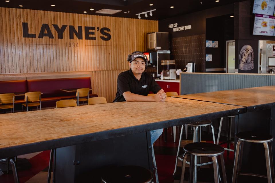 A fast food restaurant manager wears a black t-shirt and baseball cap while sitting at a high table.