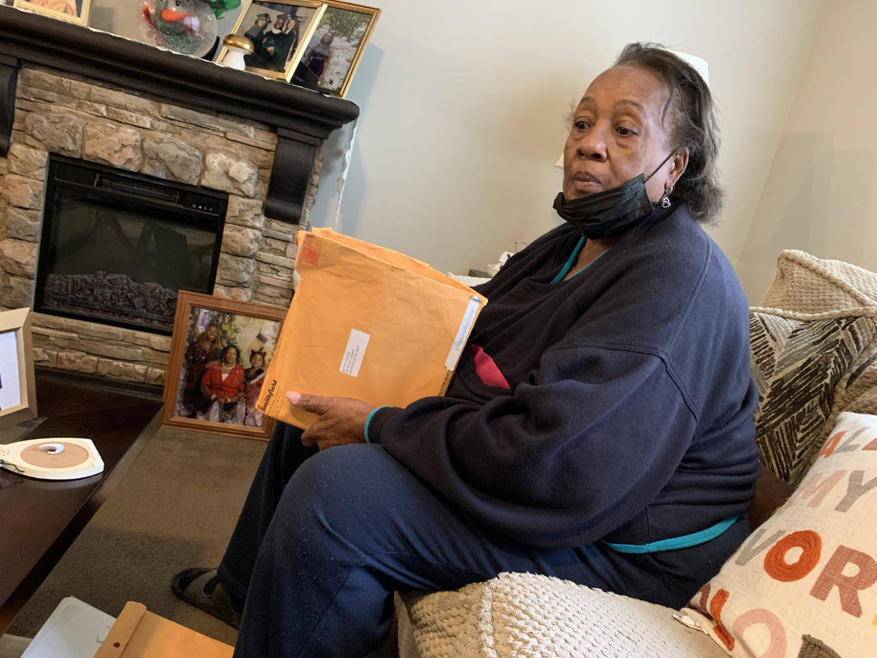 Betty Ricks sits on a couch holding an open envelope in front of a stone fireplace, around which are framed photos of people.