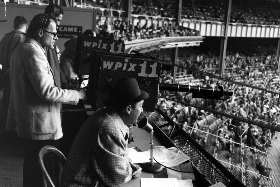 FILE - New York Yankees announcer Mel Allen, seated, reports the game between the Yankees and Baltimore Orioles from the television box behind home plate at Yankee Stadium on May 11, 1956, in New York. Many baseball fans, especially older ones, originally fell in love with America’s pastime by listening to ballgames on AM radio. But several major automakers are eliminating broadcast AM radio from newer models, prompting lawmakers on Capitol Hill to propose legislation that would prevent the practice for safety and other reasons. (AP Photo/Robert Kradin, File)