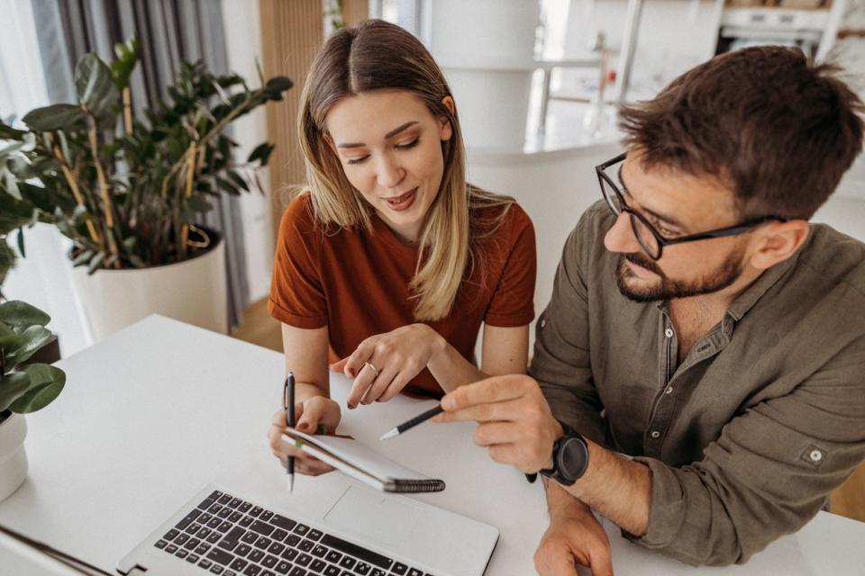 Two people sit in front of a laptop while looking at a notepad, pens in their hands. 