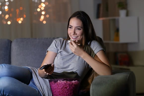 Happy young woman on the couch with a TV remote and a bucket of popcorn.