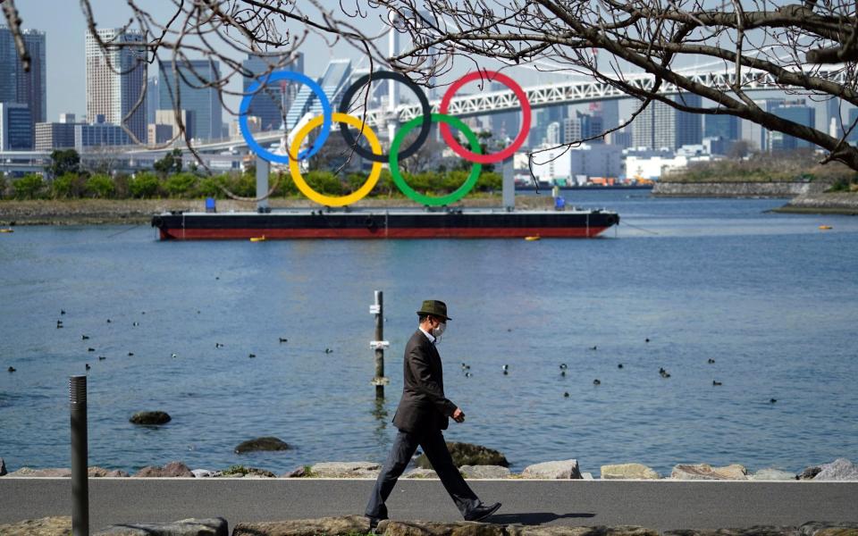 A man walks with a backdrop of the Olympic rings floating in the water in the Odaiba section in Tokyo - Eugene Hoshiko/AP