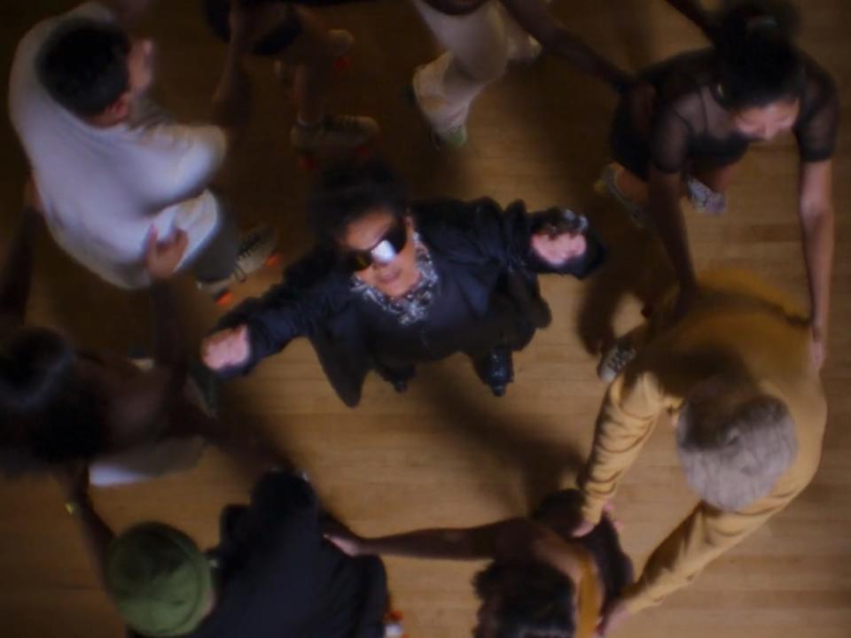 kris jenner dancing in a circle of roller skaters. she's wearing sunglasses and has hair hands in the air, and is viewed from a camera above