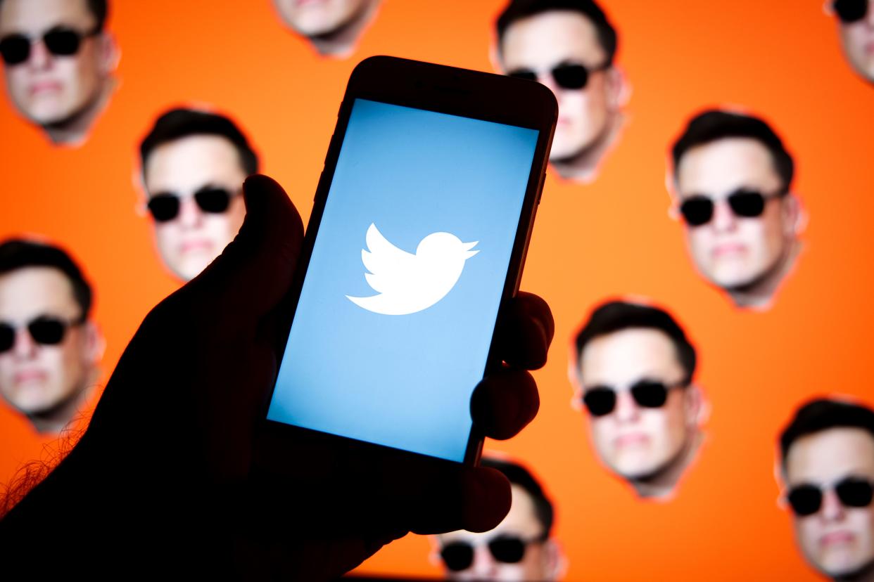 The Twitter logo is seen on a mobile device being held in front of several images of Elon Musk's face