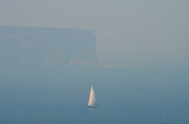 A sailing boat is seen through smoke haze from bushfires in front of the cliffs of North Head, Sydney Harbour