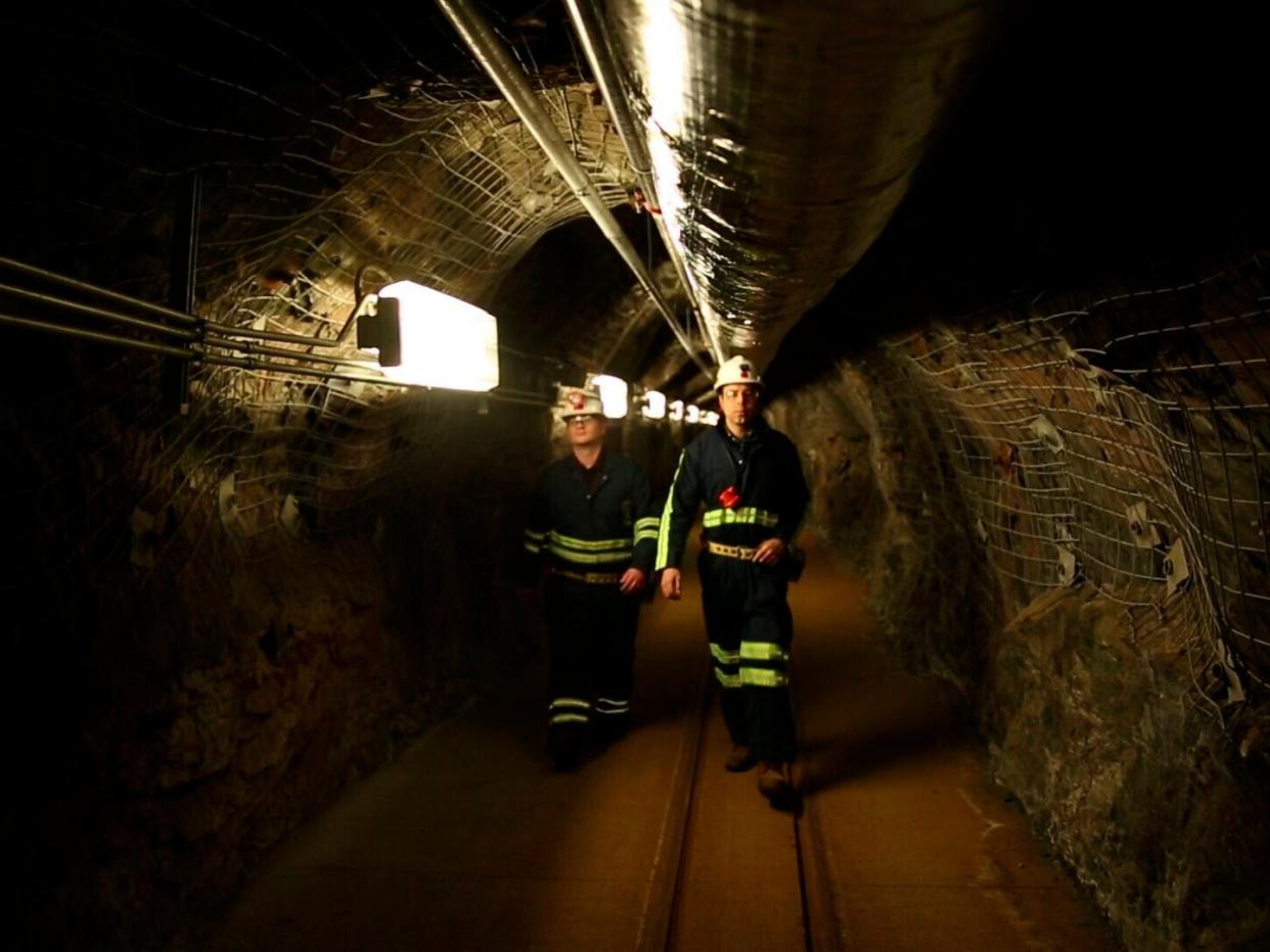 two researchers walk down mining tunnel wearing black jumpsuits with yellow stripes and white construction helmets
