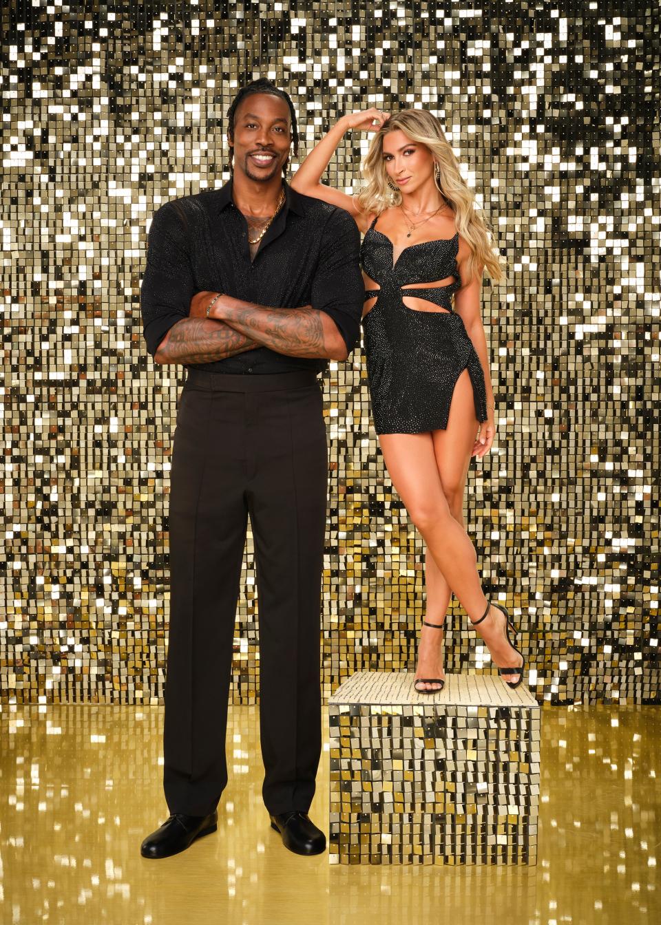 Dwight Howard stands in a black shirt and pants, while daniella karagach poses beside him on a gold platform in a black cut-out dress against a sparkly background