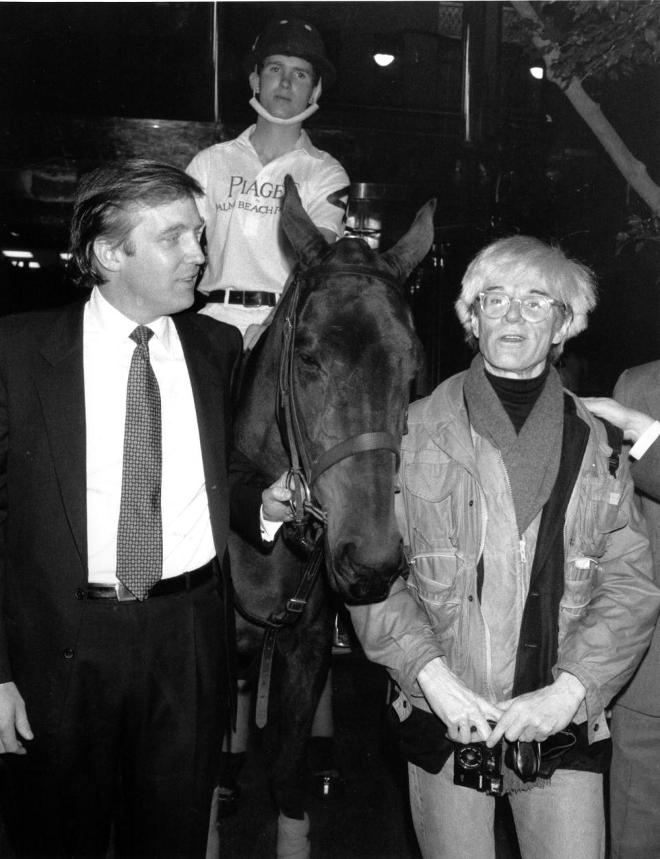 <p>Donald holds the bridle of a polo pony while talking to Andy Warhol on Nov. 4, 1983. Yale University polo player Eric Stever sits astride the horse. <i>(Photo: Mario Suriani/AP)</i> </p>