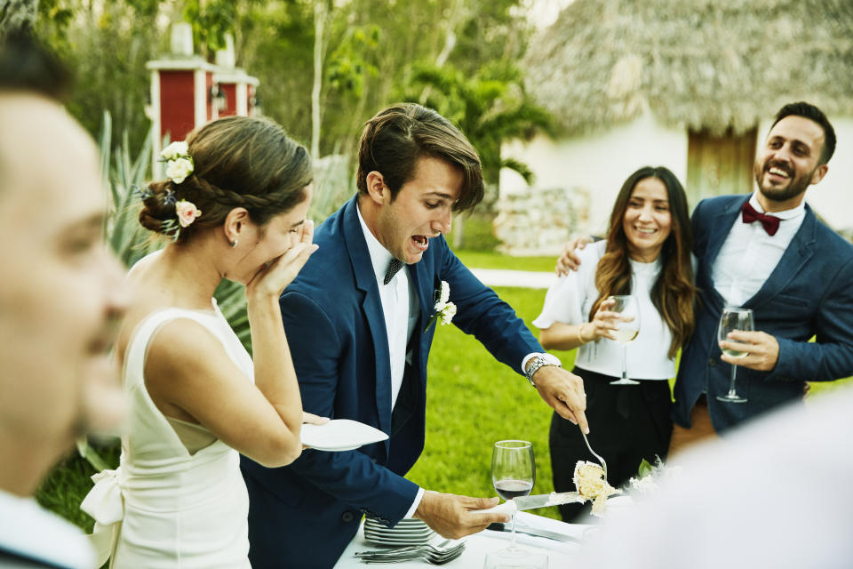 Laughing bride and groom cutting cake for guests during outdoor wedding reception