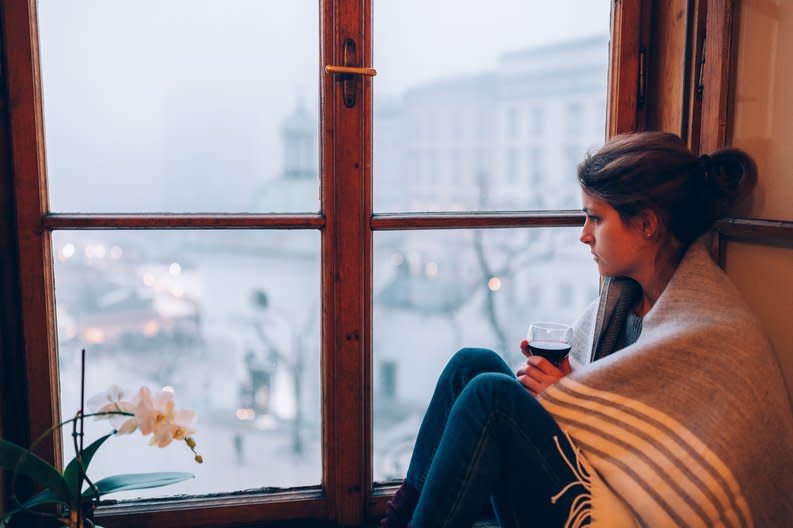 A worried woman wrapped in a blanket sitting at a window.