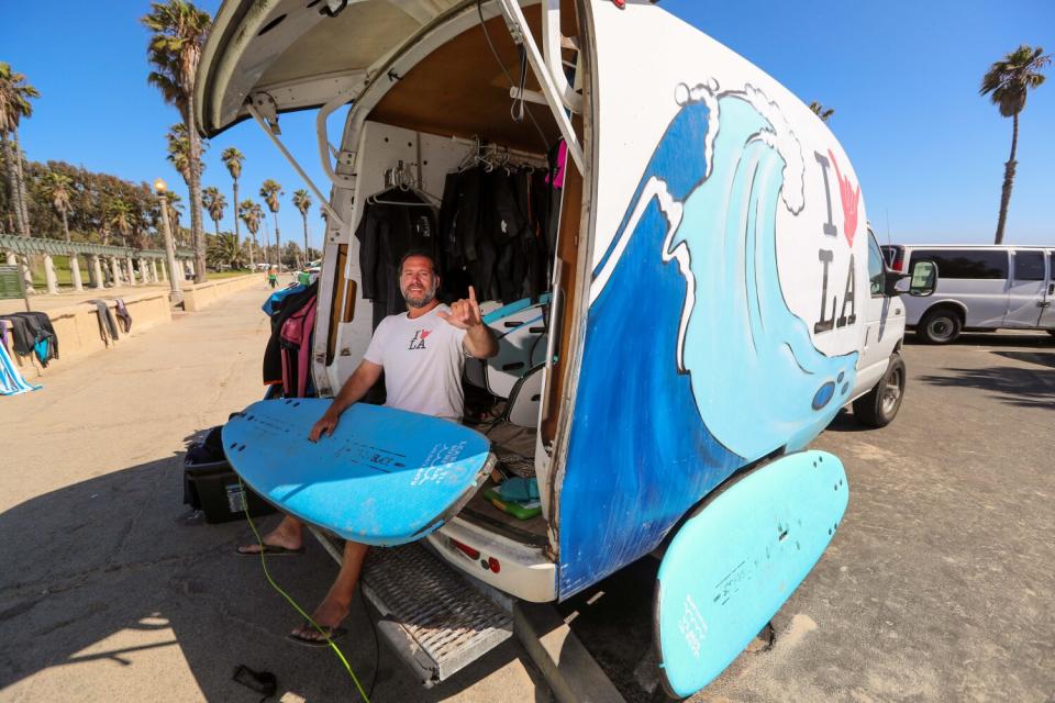 A man holding a surfboard sits in the back of a truck with surfing equipment.