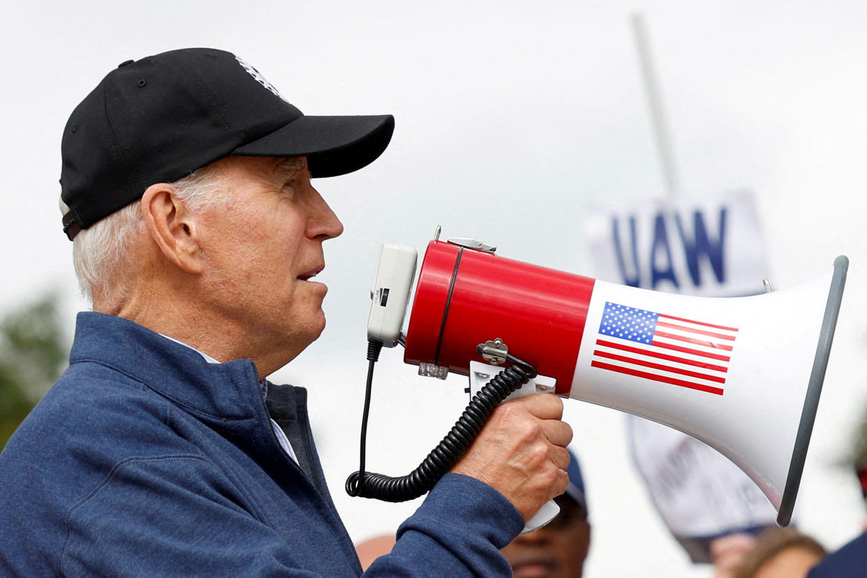 President Biden, in profile, wears a black baseball hat and holds a megaphone emblazoned with an American flag to his mouth in front of a sign reading: UAW.