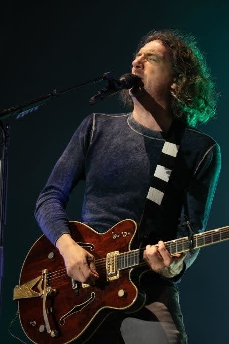 Gary Lightbody, wearing a casual long-sleeve top, plays a guitar and sings into a microphone on stage