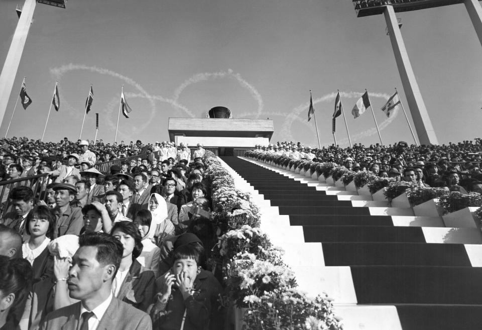 FILE - In this Oct. 10, 1964, file photo, five interlocking Olympic rings are thrown high in the sky by jet planes drift over the stadium during the opening ceremonies for the 1964 Olympics at the National Stadium in Tokyo. The famous 1964 Tokyo Olympics highlighted Japan’s resiliency. It was a prospering country that was showing off bullet trains, transistor radios, and a restored reputation just 19 years after devastating defeat in World War II. Now Japan and Tokyo are on display again, attempting to stage the postponed 2020 Tokyo Olympics in the midst of a once-in-a century pandemic. (AP Photo, File)