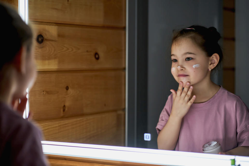 Girl applies skincare cream, looking at mirror, reflecting beauty routine