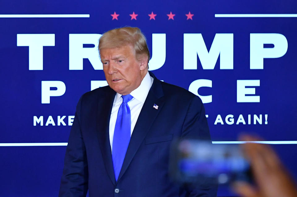 Donald Trump stands in front of a large sign reading: Trump Pence - Make America Great Again.