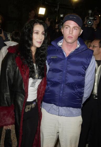 Cher in a black leather trench coat and Elijah Blue Allman are posing together in front of a group of photographers