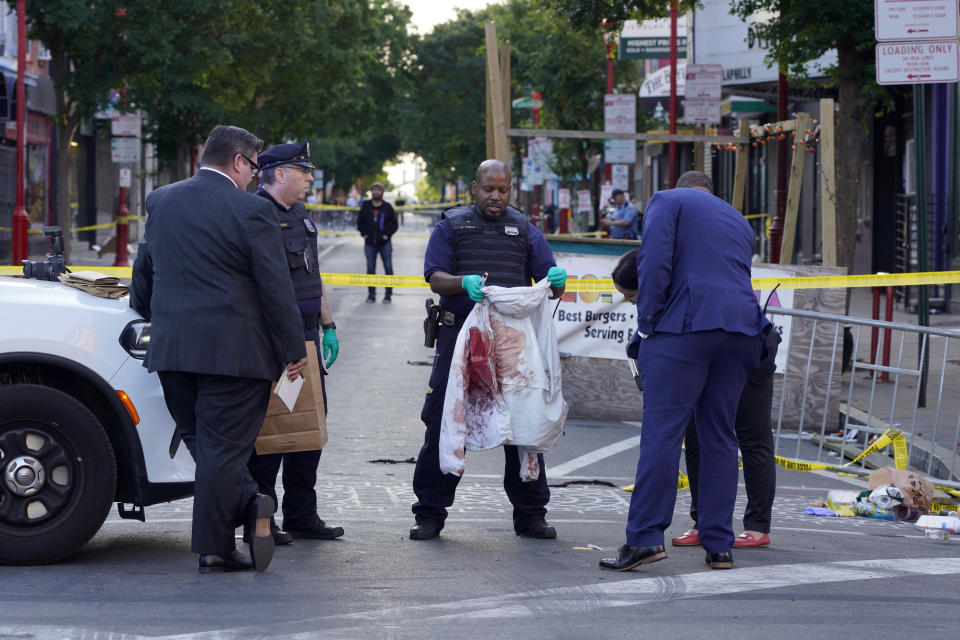 A police officer holds up a jacket that appears to have blood stains on it as other officials stand near a taped-off street.
