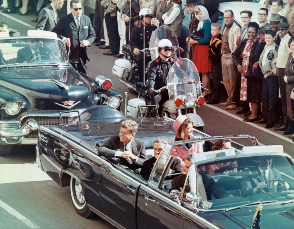 John F. Kennedy and Jacqueline Kennedy ride in a motorcade in an open-top car, with Secret Service agents and a large crowd of onlookers