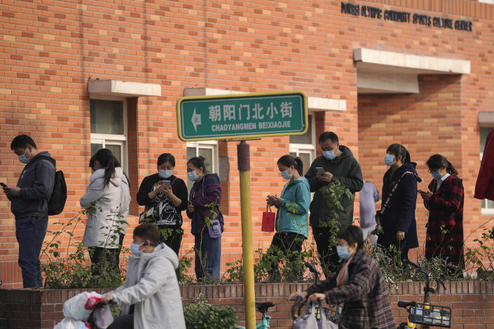 Women wearing face masks to help curb the spread of the coronavirus ride scooters passing by masked residents line up to receive booster shots against COVID-19 at a vaccination site in Beijing, Monday, Oct. 25, 2021. A northwestern Chinese province heavily dependent on tourism closed all tourist sites Monday after finding new COVID-19 cases. (AP Photo/Andy Wong)