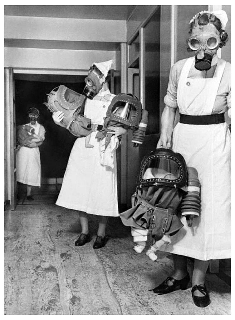 Three nurses are wearing gas masks and holding infants also in gas masks in a hospital hallway