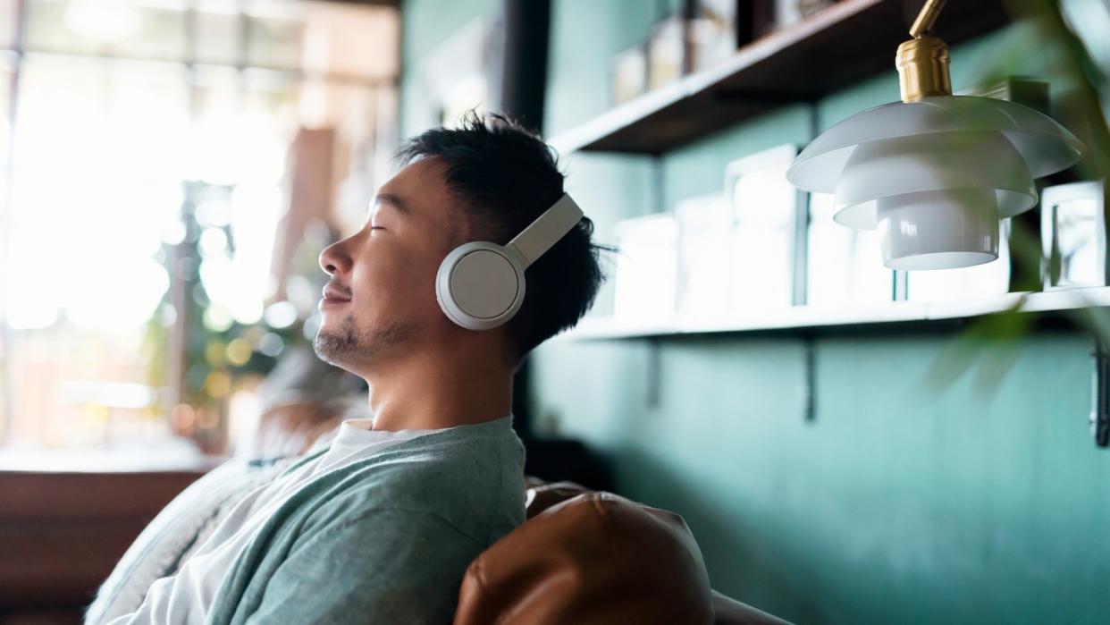 young man with eyes closed, enjoying music over headphones while relaxing on the sofa at home