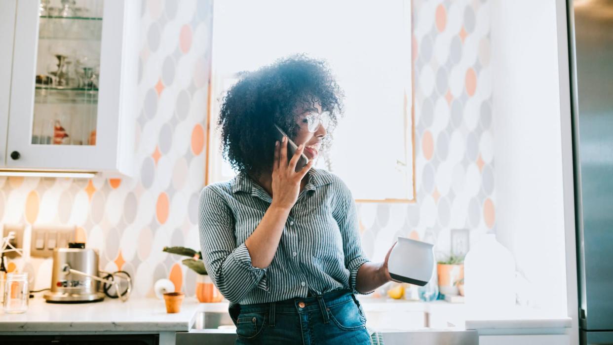 A happy young adult woman enjoys time at her home, talking on the phone in the kitchen while drinking a cup of coffee.