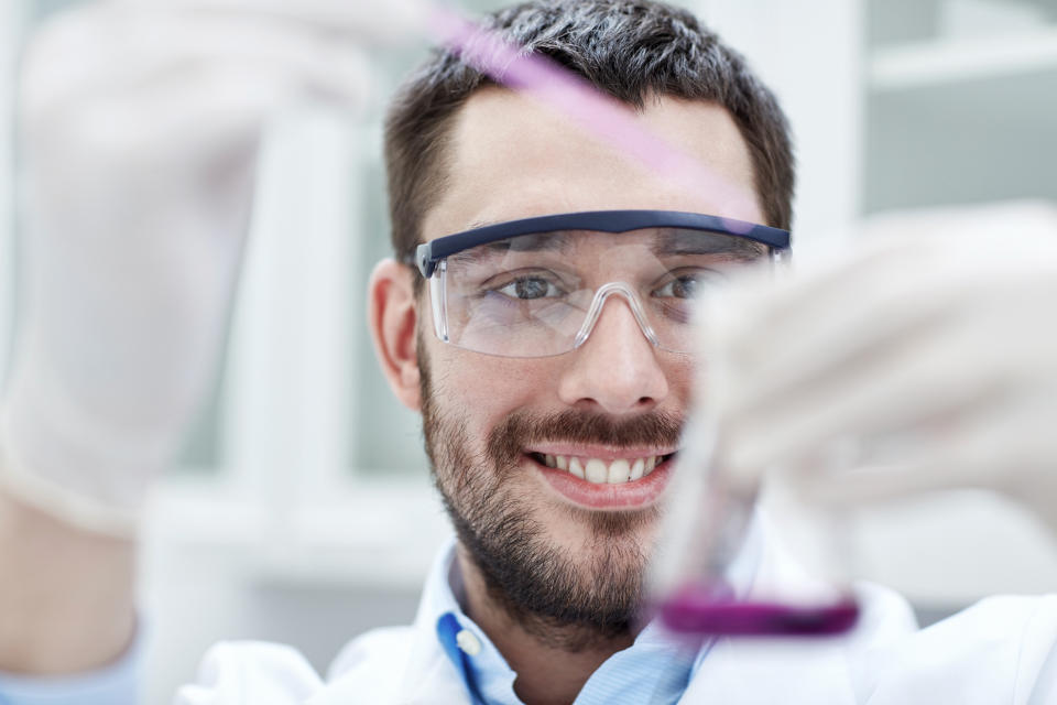 Smiling man in a lab coat using a pipette to put purple liquid into a beaker.