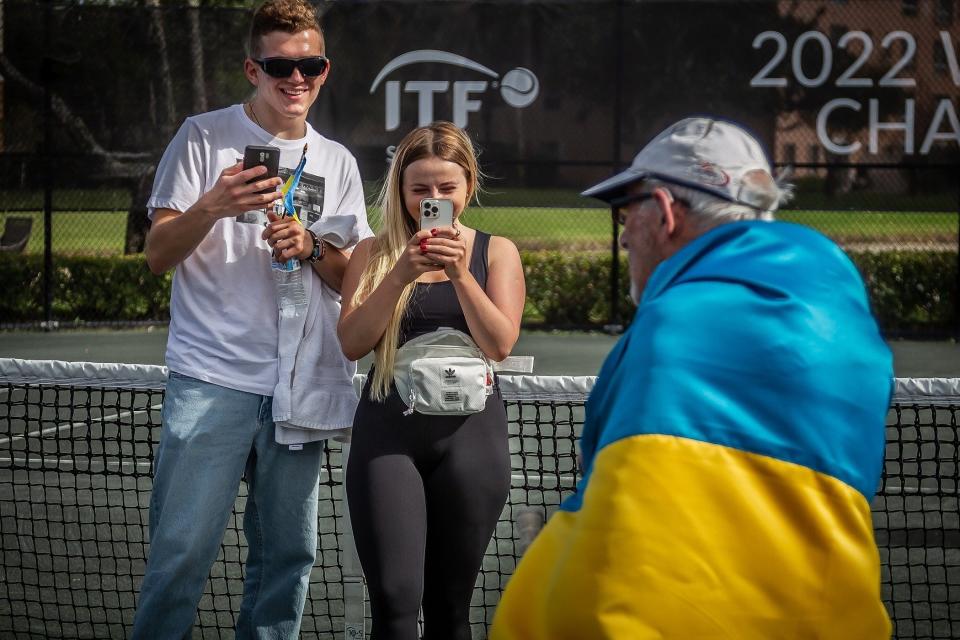 Ukrainian refugees Elister Haievskyi, left, and Katya Bodnar, center, film fellow countryman and refugee Leonid Stanislavskyi, 98, during men's singles action at the  ITF Super Seniors World Tennis Championships at Coral Lakes Tennis Center in Boynton Beach, Monday. Stanislavskyi is competing in the over-90 division of the tournament.