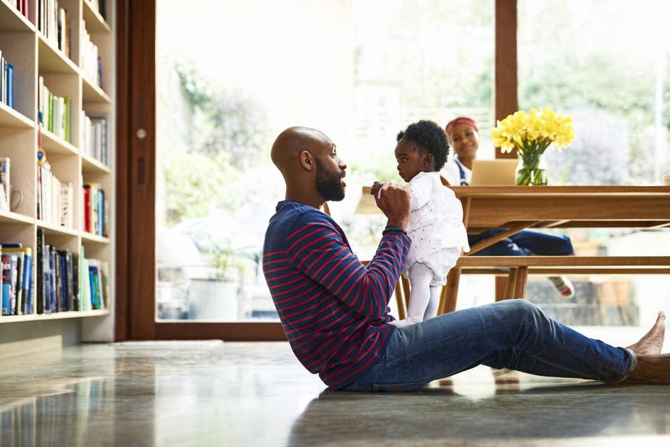 Father playing with baby daughter (6-11 months), woman using laptop at table in background