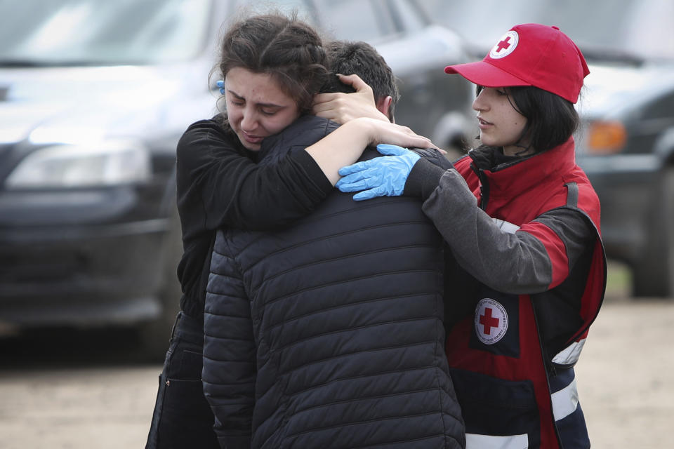 Two people embrace while someone wearing a red hat and vest, each emblazoned with a red cross, holds a gloved hand on the back of one of them.