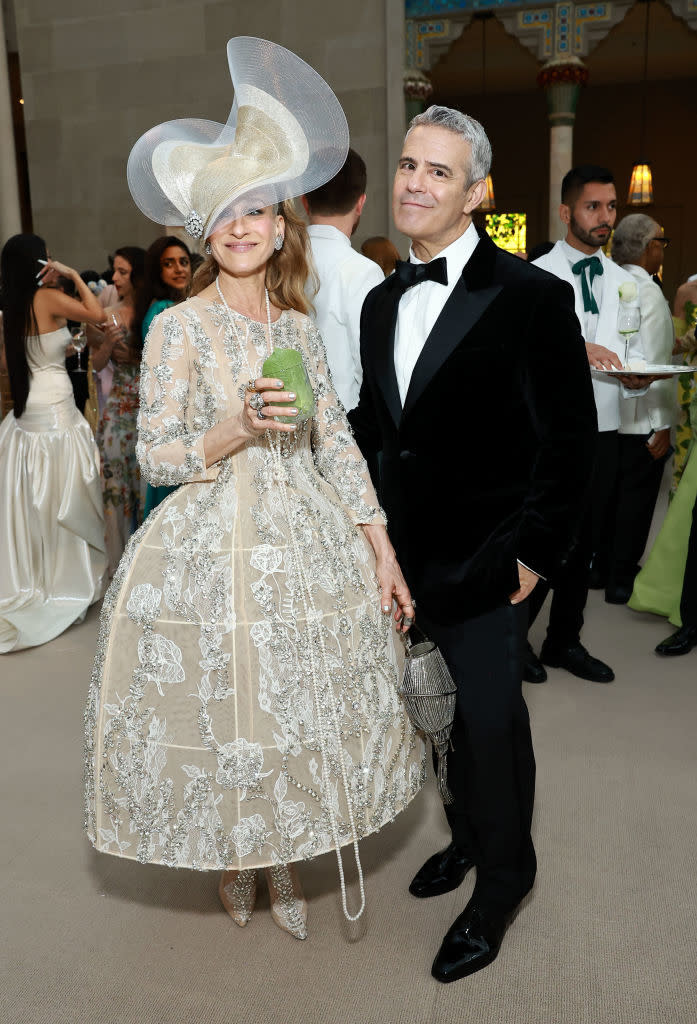 Two individuals at an event, one in a lace dress with a large hat and the other in a black tuxedo