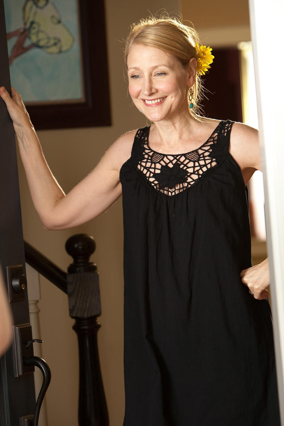 Patricia Clarkson in a black sleeveless dress with lace detail, smiling with a flower in her hair, indoors
