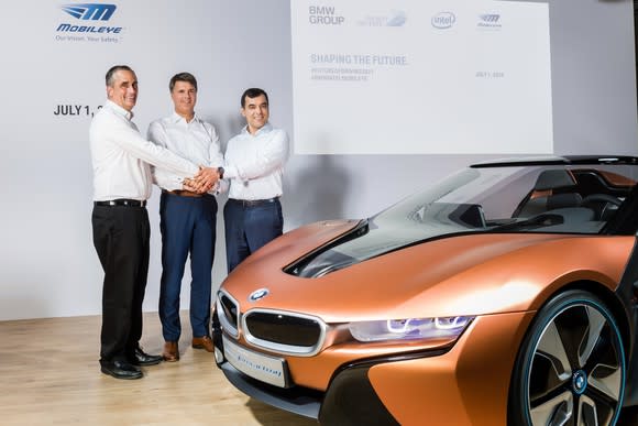 The three executives are standing on a stage next to a BMW i8 hybrid sports car.