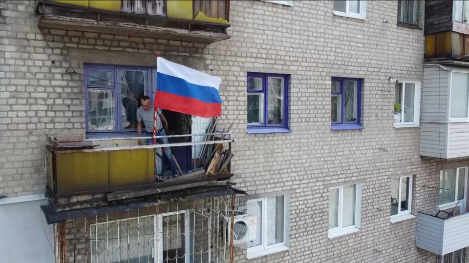 A man stands on a dilapidated balcony of a brick building next to a Russian flag.