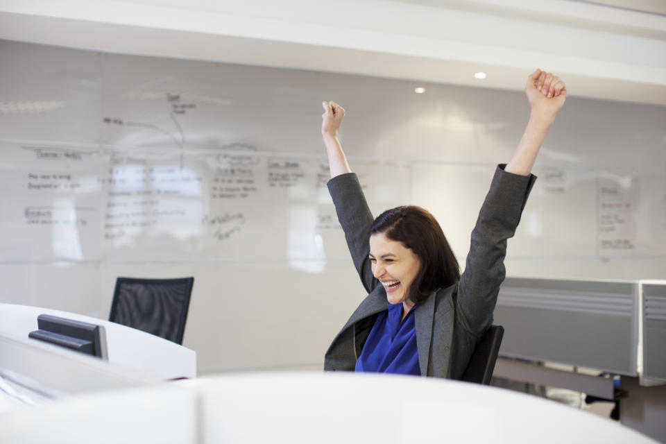 A woman in a suit throws her hands in the air in victory looking at her computer in an office.