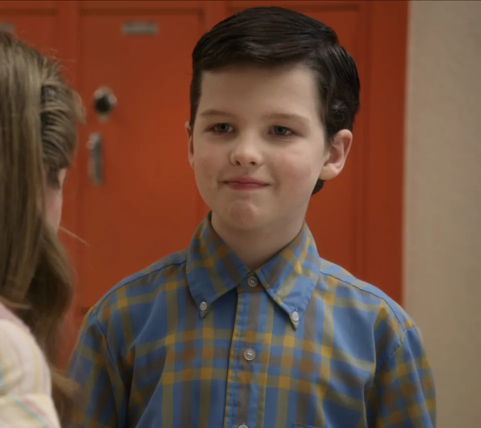 Young Sheldon in a plaid shirt standing in front of lockers