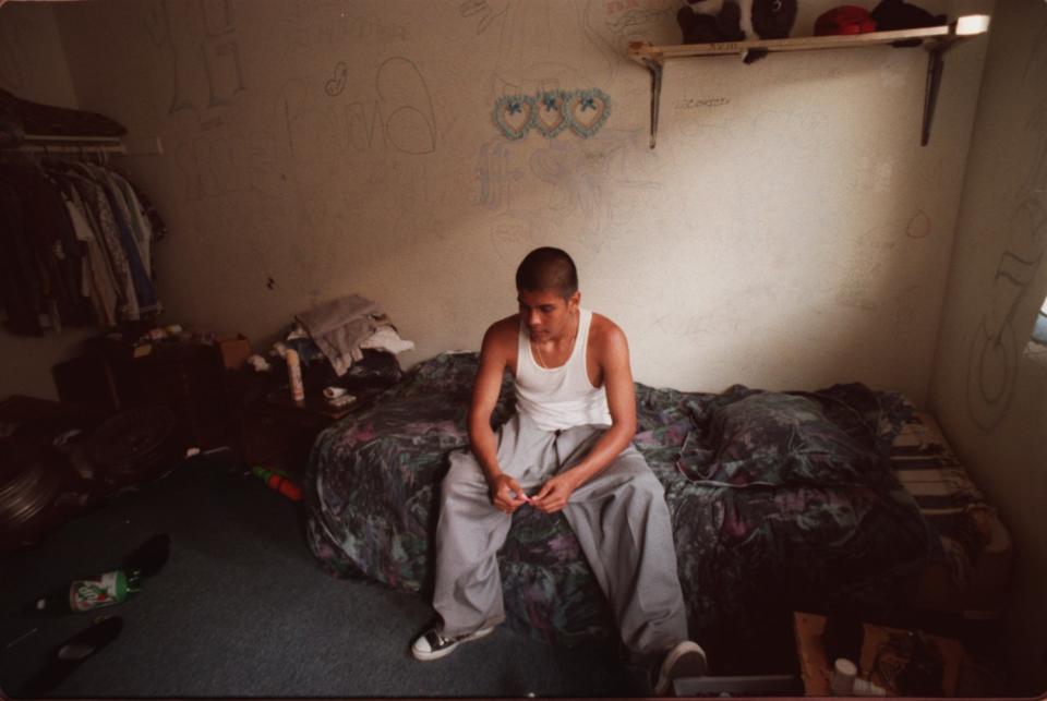 An 18th Street gang member sits on a twin bed in a small, graffiti-covered bedroom.