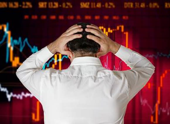 A man holds his head while looking at a screen with a chart of a declining stock price