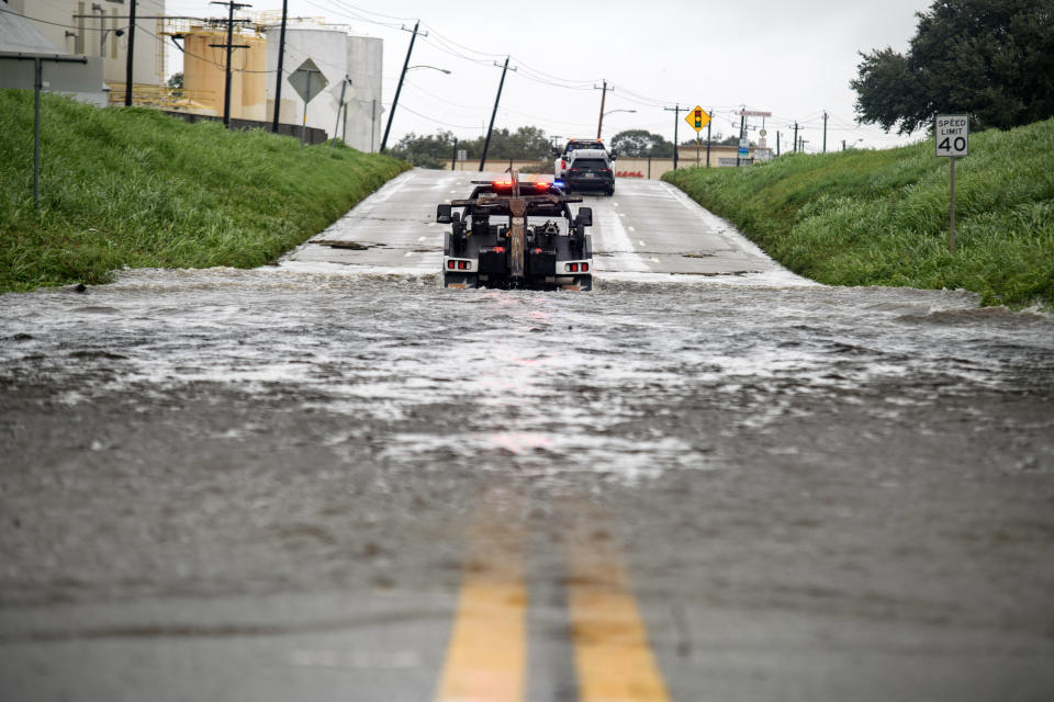 A police vehicle with flashing lights is partially submerged in floodwater blocking a road. Surrounding area is wet and there are traffic signs and trees in the background