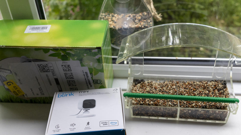 A Blink Mini box, plastic bird feeder with seeds in on a windowsill