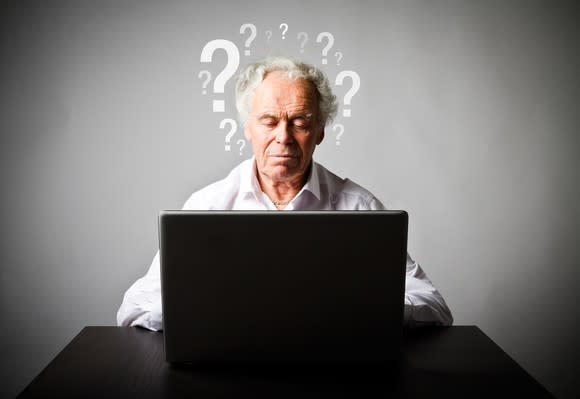 A retiree in front of a laptop computer with question marks on a wall behind him.