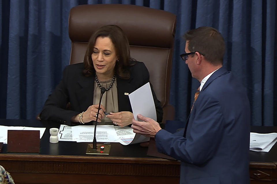 Vice President Kamala Harris seated at the microphone in her presiding chair at the U.S. Senate, with an aide carrying a document to her left.