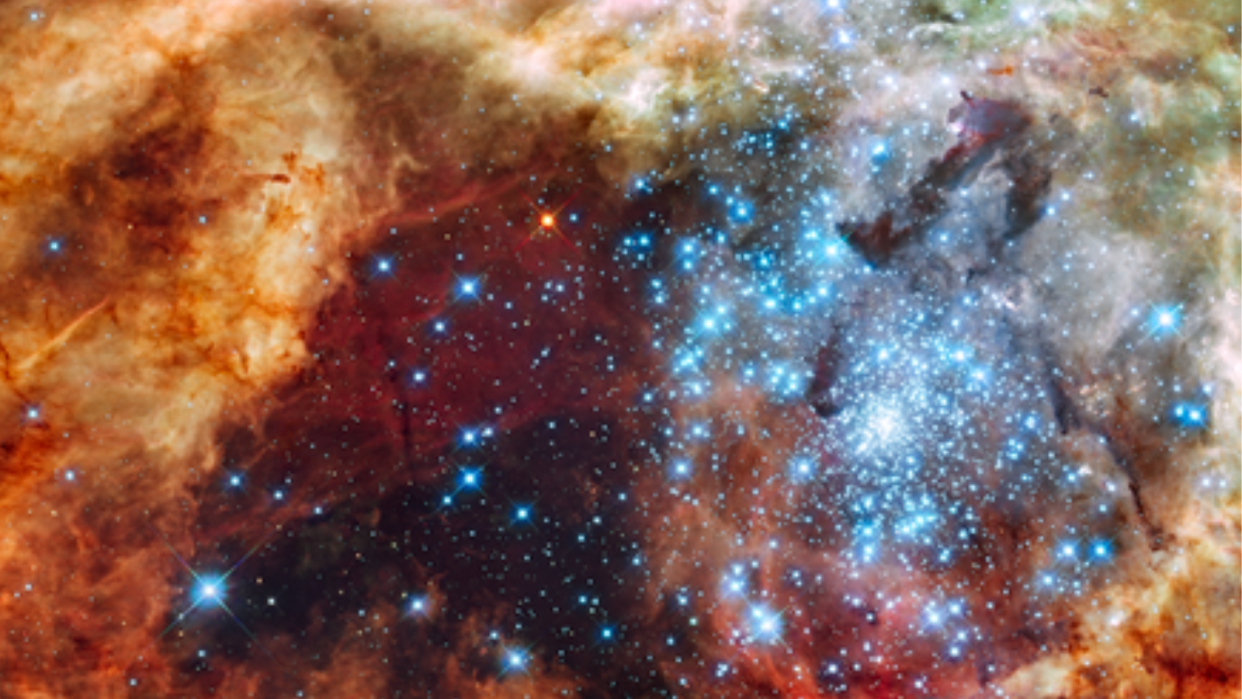  A hazy scene with orange and burgundy gaseous clouds. A burst of bright blue stars is concentrated toward the right. A single red star is seen in the center-top. 