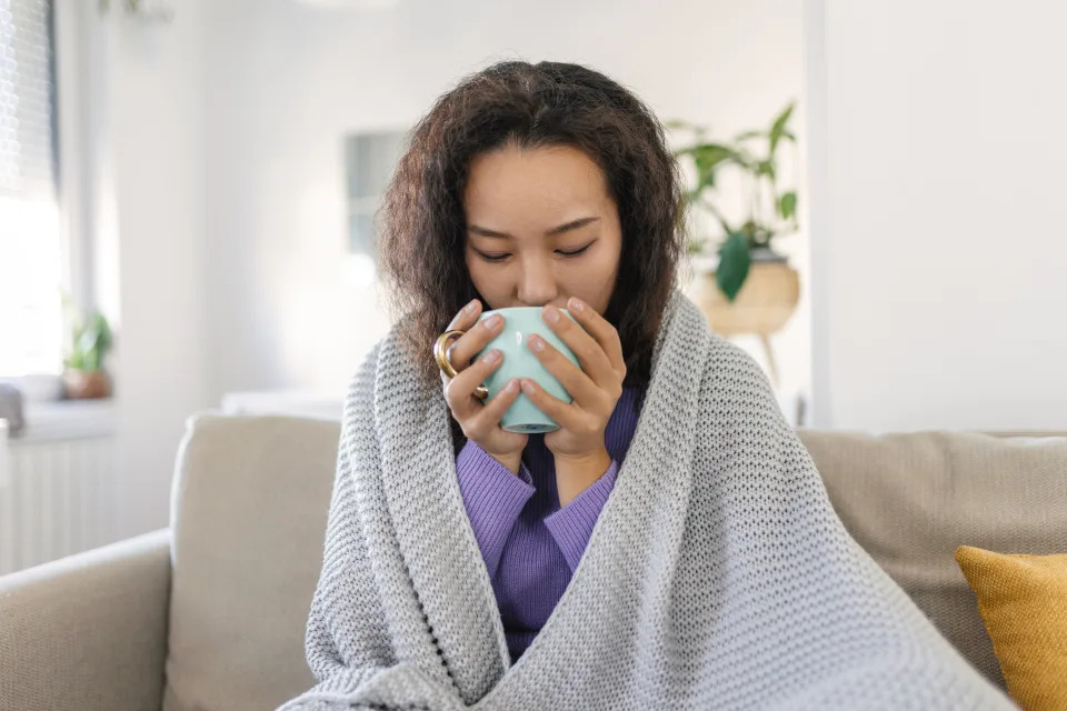 Wanita muda Asia yang sakit duduk di rumah di tempat tidur dengan secangkir teh hangat dan sapu tangan. Pilek musiman, batuk, pilek, infeksi virus, pengobatan di rumah