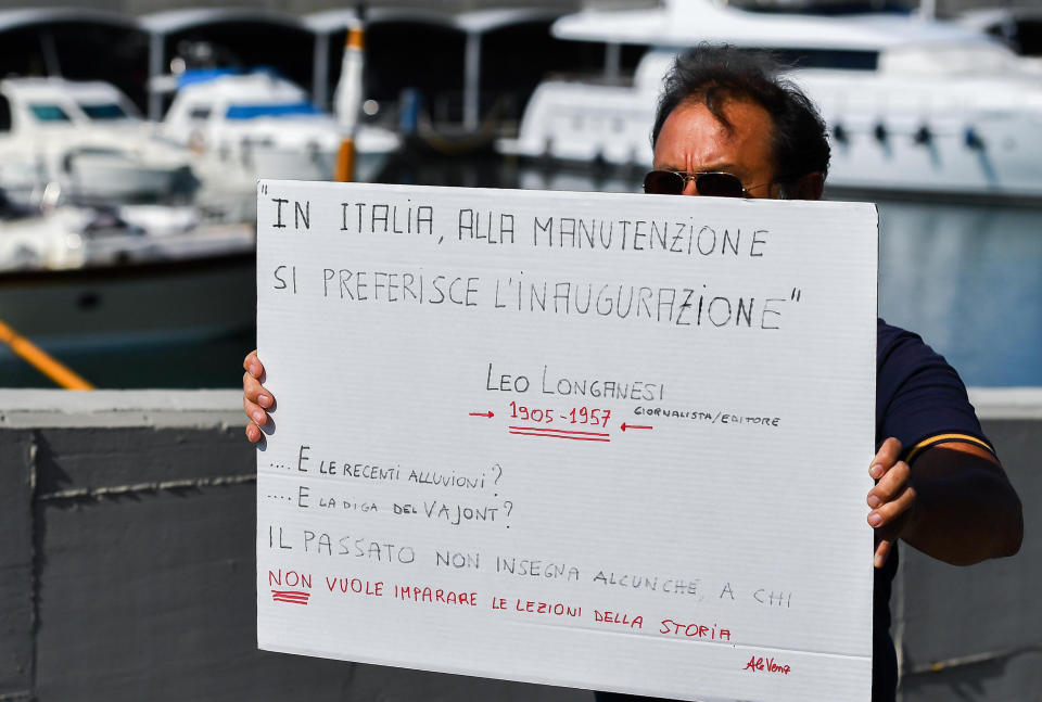 A man holds up a sign with writing reading in Italian "In Italy, at maintenance it is preferred inauguration" and the recent floods? and the Vajont dam? The past doesn't teach anything to those who don't want to learn the lessons of history" as he stands outside the exhibition center Fiera di Genova where a funeral service for some of the victims of a collapsed highway bridge is taking place, in Genoa, Italy, Saturday, Aug. 18, 2018. Saturday has been declared a national day of mourning in Italy and includes a state funeral at the industrial port city's fair grounds for those who plunged to their deaths as the 45-meter (150-foot) tall Morandi Bridge gave way Tuesday. (Simone Arveda/ANSA via AP)