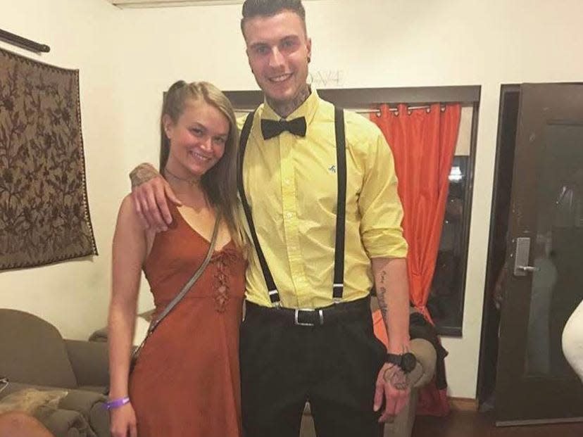 Photo of Emily May and her boyfriend Jakob. Emily has long blonde hair and wears an orange sleeveless dress with a lace up front and black platform sandals. Jakob had dark brown hair and tattoos on his hands, left arm, and neck. He wears a yellow button-down shirt, black bow-tie, suspenders, black pants, and black shiny shoes.